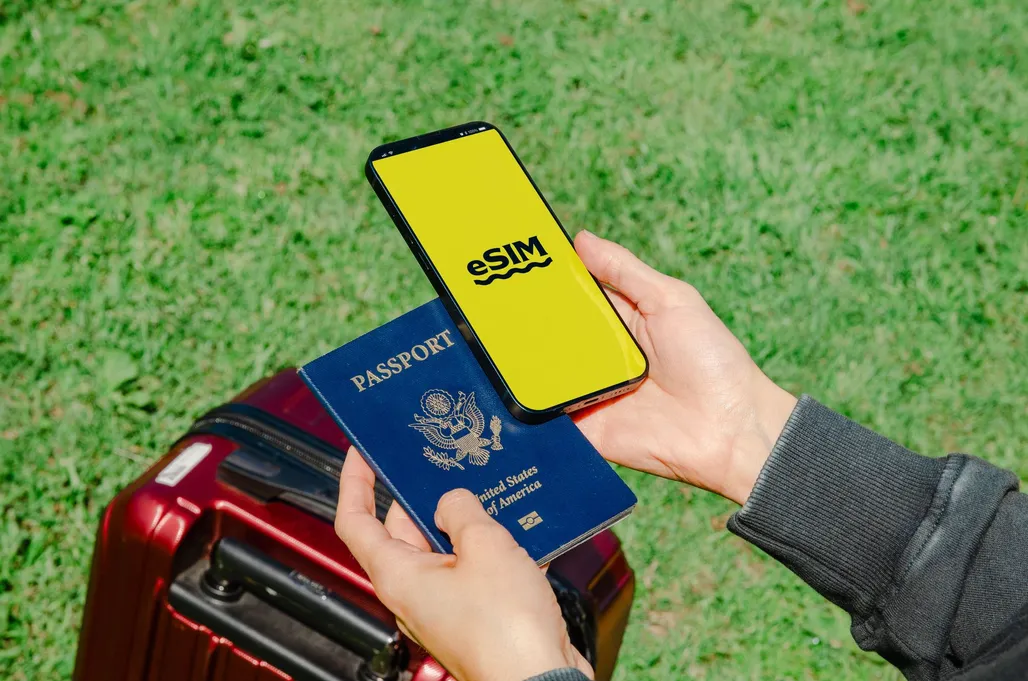 ידיים מחזיקות סמארטפון עם מסך eSIM לצד דרכון ומזוודה